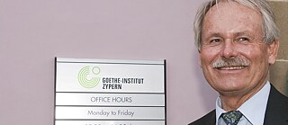 A sign with the logo and the opening times of the Goethe-Institut Cyprus hanging on a wall. Next to it is a man with gray hair and a mustache. He is wearing a suit and tie and smiles while gazing to the left.