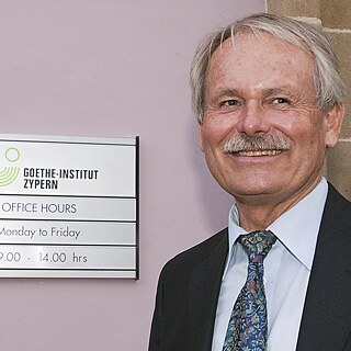 A sign with the logo and the opening times of the Goethe-Institut Cyprus hanging on a wall. Next to it is a man with gray hair and a mustache. He is wearing a suit and tie and smiles while gazing to the left.