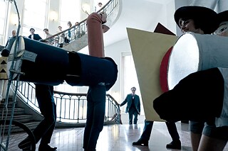 Standbild aus der Serie “Die neue Zeit“: Ein buntes Treiben von  Studenten, darunter einige in Kostümen, im Bauhaus Treppenhaus, Itten und Gropius sind in der Menge zu sehen.