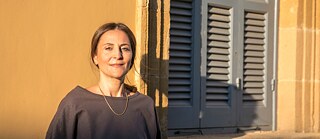 A lady stands smiling in front of a yellow ocher wall with a blue wooden window, under the afternoon sunlight. She has her long brown hair tied, her hands in both pockets of her black trousers and she wears a grey blouse and a neck chain.