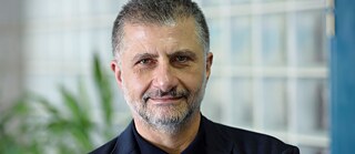 Half portrait of a middle-aged man in a black suit. He has short black and gray hair, a gray beard, brown eyes, and smiles at the camera. In the background you can see a blurred plant.