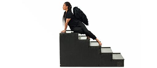 The artist leaning against black stairs wearing a black costume with wings