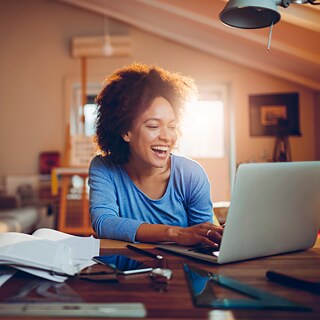 Eine junge Frau arbeitet an einem Laptop. Auf dem Tisch liegen Bücher, ein Handy und Notizen. Im Hintergrund sind Bückerregale, eine Couch und weitere Einrichtungsgegenstände unscharf zu sehen.