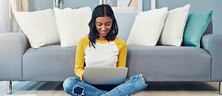 Eine junge Frau in modischen Jeans sitzt im Wohnzimmer auf dem Boden vor einer Couch und blickt lächeln auf ein Laptop.