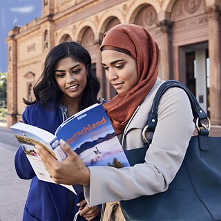 Zwei Frauen stehen mit einem Lehrbuch vor einem Gebäude und schauen interessiert ins Buch