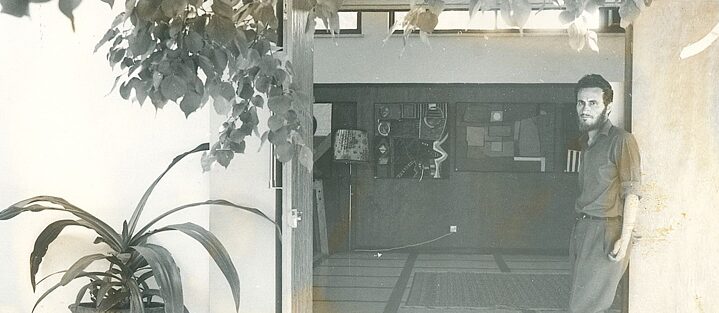 Christoforos Savva steht auf der rechten Seite des Eingangs seiner Ausstellung und schaut in die Kamera. Er trägt Anzughosen, ein Hemd und schwarze Schuhe, seine Arme hängen locker nach unten. Seine Haare sind dunkel und er trägt einen Vollbart. Einige Bilder der Ausstellung die auf der Wand hängen sind vage durch die offene Tür zu erkennen. Links neben der Tür und über der Tür sind Pflanzen.