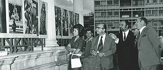 Vier Männer und eine Frau, die eine weiße Handtasche trägt, stehen im Vordergrund und betrachten Fotografien bei der „Weltausstellung der Fotografie“ (1970). Im Hintergrund stehen weitere Besucher*innen, die elegante Kleidung tragen. 