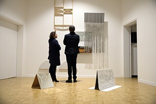 Ein Mann und eine Frau tragen elegante Kleidung und stehen mit dem Rücken zur Kamera und betrachten die Kunstwerke die an der weißen Wand hängen. Auf dem Holzboden stehen zwei weitere Kunstwerke. Auf der rechten Seite steht die weiße Tür zum Nebenraum offen.