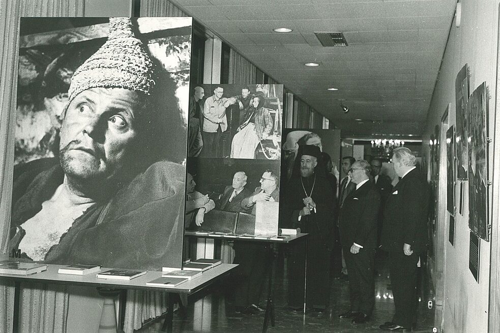The German ambassador is seen in a group with three men, with other visitors to the Brecht exhibition in the background. The men are wearing elegant suits, to the left is the guest of honour Archbishop and President of the Republic of Cyprus Makarios. The men are standing among the exhibits. In the foreground is an exhibit showing a frightened man. Below these pictures is a shelf with Brecht's literary works.