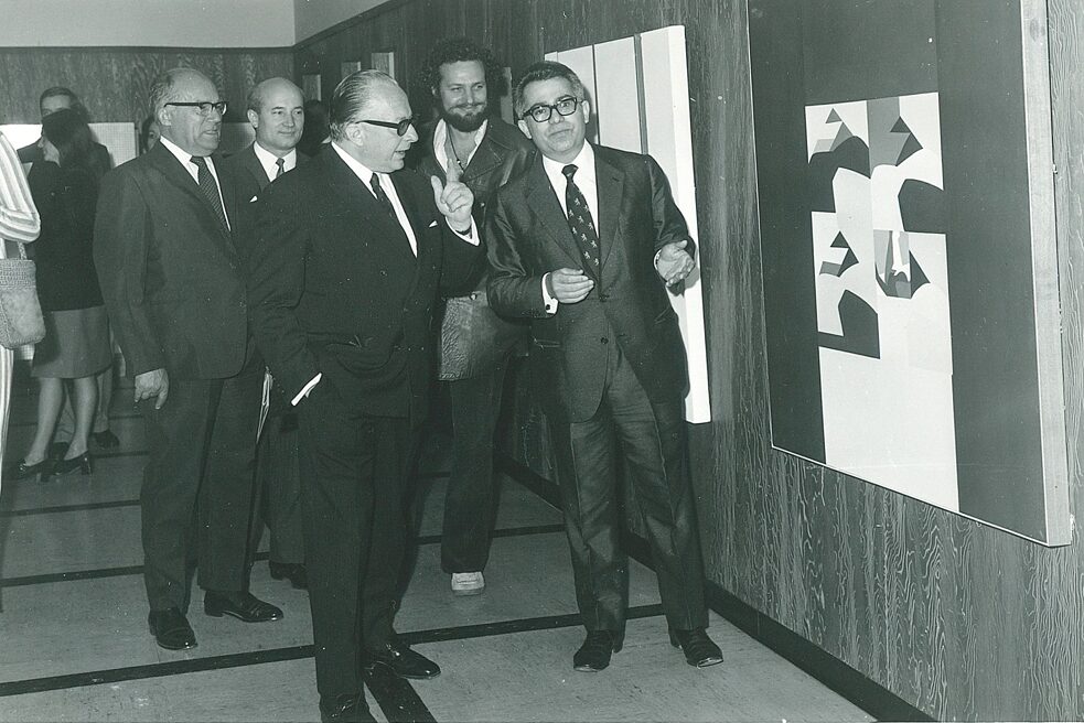 Fünf Männer im Anzug diskutieren ein abstraktes Kunstwerk bei der Ausstellung von Nikos Kouroussis 1971, welches auf der rechten Seite des Bildes auf einer Holzwand hängt. Im Hintergrund sind weitere Bilder erkennbar, welche ebenfalls an der Wand hängen. Auch dort stehen weitere Besucher*innen.