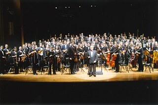 A whole orchestra stands on a lit stage, facing the camera towards the audience. The back and foreground are completely black. Another spotlight is directed at the conductor in the centre of the stage. The men and women are all standing and wearing black and white evening dress, they are uniformly dressed.