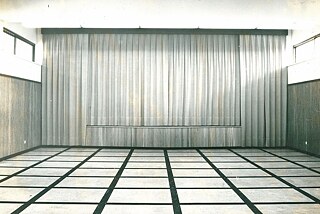 Der Saal im Goethe-Institut ohne Ausstellungsstücke. Im Fokus ist der verschlossene Vorhang der Bühne. Links und rechts sind Fenster zu sehen, auf dem Boden ist ein Muster mit hellfarbigen Quadraten und schwarzen Rändern.
