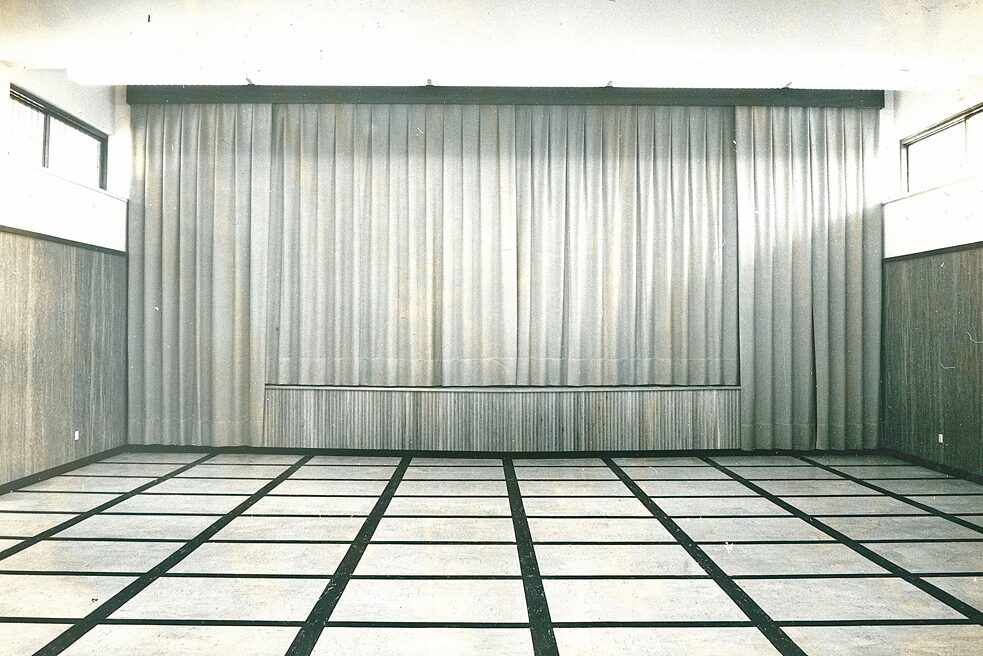 Der Saal im Goethe-Institut ohne Ausstellungsstücke. Im Fokus ist der verschlossene Vorhang der Bühne. Links und rechts sind Fenster zu sehen, auf dem Boden ist ein Muster mit hellfarbigen Quadraten und schwarzen Rändern.