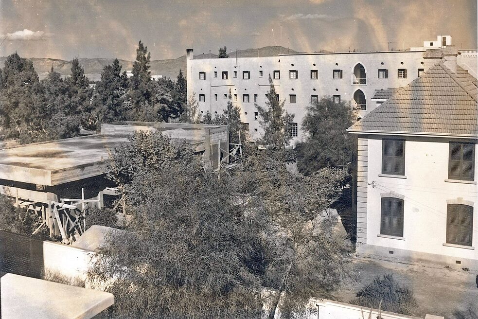 The photograph shows a larger section of the area around the Goethe-Institut building site. In the background you can see mountains and trees and the Ledra Palace Hotel. The Goethe-Institut building can be seen on the right. The construction site is surrounded by scaffolding, trees and a wall.