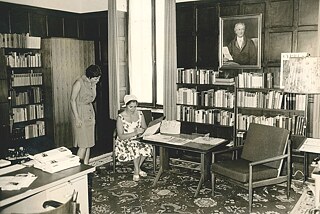 Two women are in a room, to their left is a closed window. On the right is a bookshelf, above which hangs a painted portrait of Goethe. In front of the portrait is a table with a chair on the left. A woman is sitting on it, wearing a dress and a headdress and is leafing through a folder. Another woman stands to her left and looks into the folder. Behind her is another bookcase. On the left is a desk on which office materials are placed.
