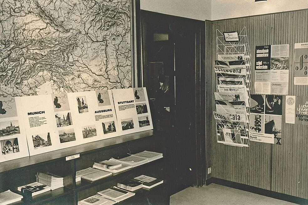 The picture shows a section of the foyer of the Goethe-Institut. On the left of the picture hangs a large map. In front of it is a shelf with books and brochures, and there are also information boards about German cities on the shelf. On the wall to the right are posters and a rack with newspapers.