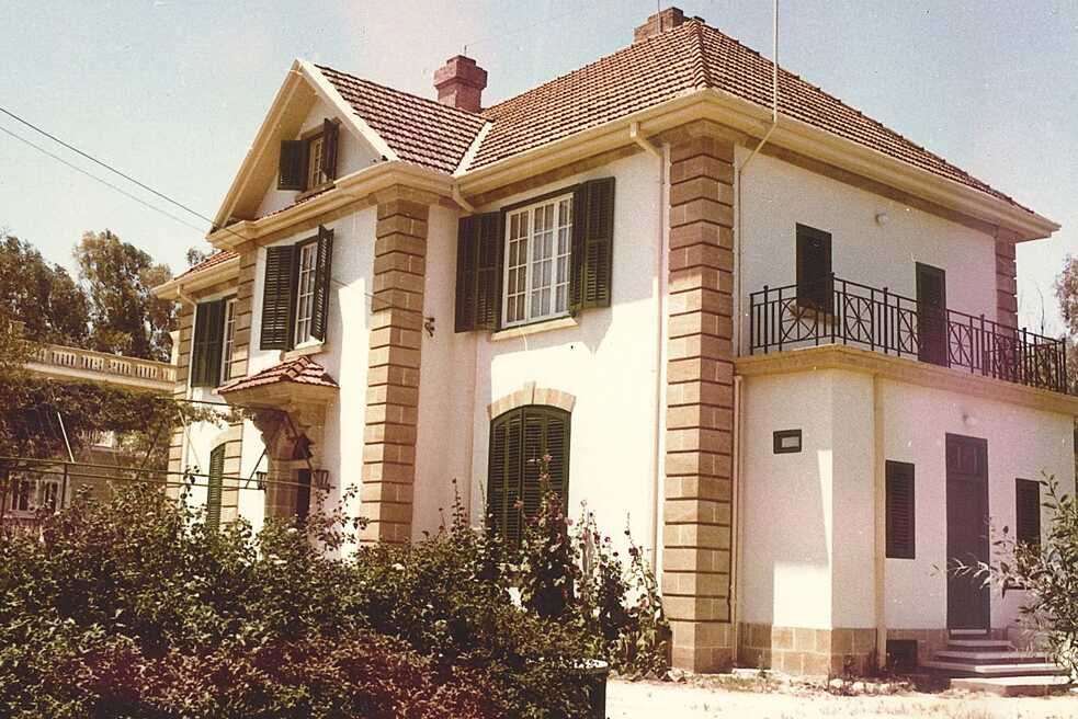 Το κτίριο του Ινστιτούτου Γκαίτε κάτω από έναν καταγάλανο ουρανό. Ο τοίχος του κτιρίου είναι άσπρος με περίγραμμα από πουρόπετρα στις γωνίες. Τα παραθυρόφυλλα είναι σκούρο πράσινο. Στα αριστερά στην άκρη της εικόνας φαίνεται ένας ανθισμένος θάμνος με μωβ άνθη.