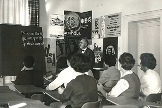 A man in a suit stands in front of a wall of posters in a classroom. On the right side of the picture is a white door. The man and his audience are looking at the blackboard. On the blackboard is written a German saying: "Drink what is clear, eat what is cooked, speak what is true!"