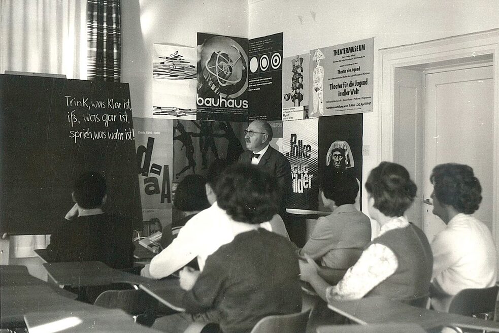A man in a suit stands in front of a wall of posters in a classroom. On the right side of the picture is a white door. The man and his audience are looking at the blackboard. On the blackboard is written a German saying: "Drink what is clear, eat what is cooked, speak what is true!"