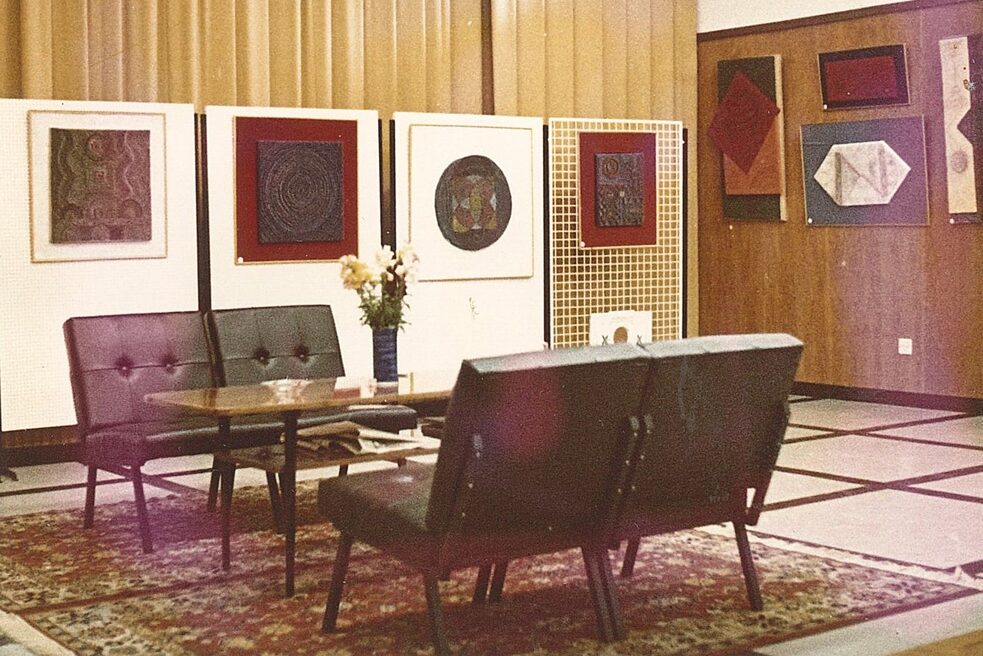 On the left side there are four pinboards on which four works of art are hanging. Behind the pinboards hang yellow curtains. On the right side, four more artworks hang on the wooden wall. On a red-beige carpet in the foreground and in the middle of the room, a table and four black armchairs are placed, on the table flowers are arranged decoratively.