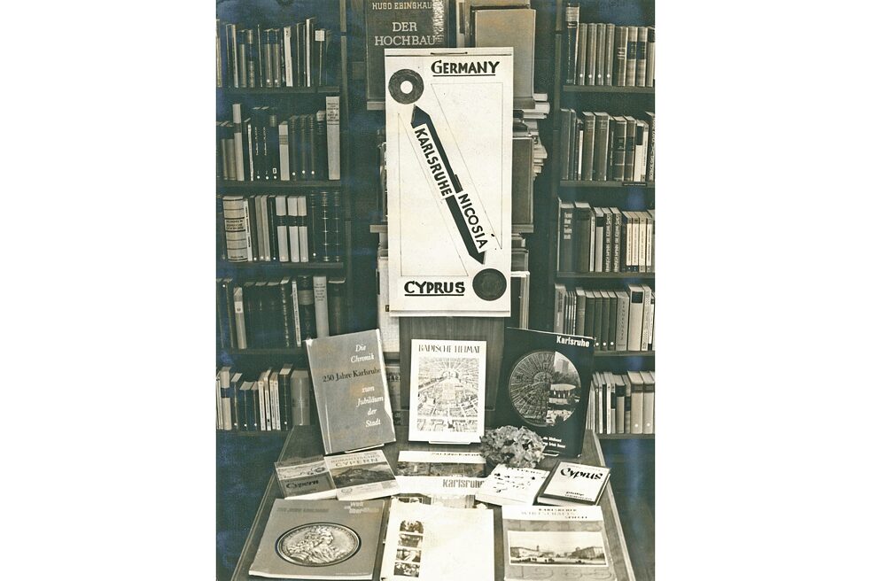 A table has been set up in front of a bookshelf for the Karlsruhe-Nicosia town-twinning. On the table are various books on the subject. Above it hangs a poster on which a connection between Karlsruhe and Nicosia as well as Germany and Cyprus is illustrated by arrows.