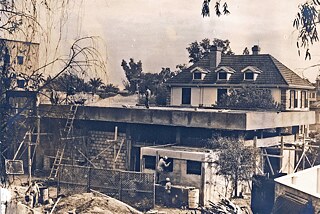 The picture shows the event hall of the institute under construction. The unfinished building is surrounded by ladders and scaffolding. A man is standing on the roof, other workers are at the bottom of the building and only their backs can be seen. At the edge of the picture are plants and trees framing the image. On the right side behind the construction site, the building of the Goethe-Institut can be seen. On the left side, part of the Ledra Palace Hotel is visible.
