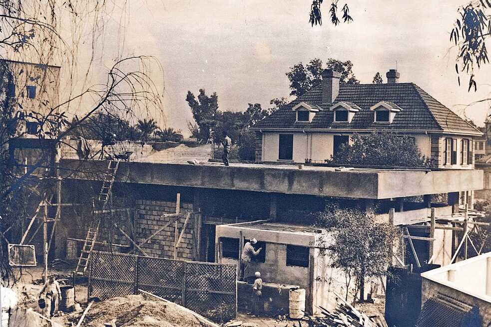 Η εικόνα δείχνει την αίθουσα εκδηλώσεων του ινστιτούτου υπό κατασκευή. Το ημιτελές κτίριο περιβάλλεται από σκάλες και σκαλωσιές. Ένας άντρας στέκεται στη στέγη, άλλοι εργάτες βρίσκονται στο κάτω μέρος του κτιρίου και μόνο οι πλάτες τους είναι ορατές. Στην άκρη της εικόνας βρίσκονται φυτά και δέντρα που πλαισιώνουν την εικόνα. Το κτίριο του Ινστιτούτου Γκαίτε διακρίνεται στη δεξιά πλευρά πίσω από το εργοτάξιο. Μέρος του Ξενοδοχείου Λήδρα Πάλας φαίνεται στα αριστερά.