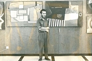 Christoforos Savva stands with his back to two abstract paintings in his exhibition. He is wearing suit trousers, a shirt and black shoes, his arms are folded. His hair is dark and he has a full beard.