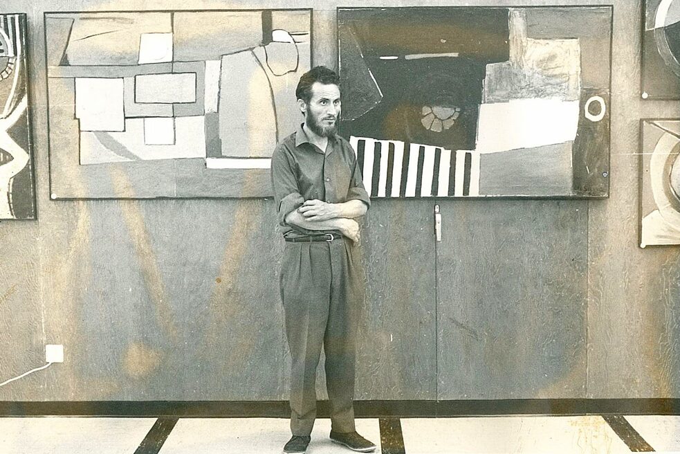 Christoforos Savva steht mit dem Rücken zu zwei abstrakten Gemälden seiner Ausstellung. Er trägt eine Anzughose, ein Hemd und schwarze Schuhe, die Arme sind verschränkt. Sein Haar ist dunkel und er hat einen Vollbart.