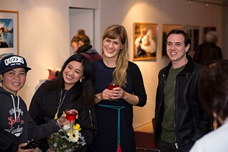 Three women and a man are standing together laughing and looking to the left. The woman on the left is wearing a peaked cap, a dark blue lettered hoodie and holding a red drink. The second woman from the left is wearing black clothes and a long necklace, in front of her is a bouquet of yellow flowers. The second woman from the right has blond hair and fringes, her dress is dark blue. She is holding a red drink with both hands. On the right is a man wearing a leather jacket. A shoulder in a white top can be seen in the front right. Individual people can be seen in the background looking at artworks in the exhibition. 