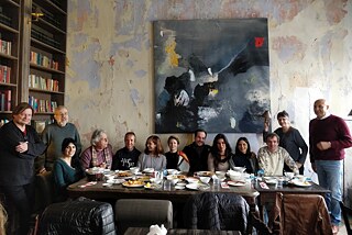 Many people are sitting at a large, set table, looking at the camera. On the left side is a full bookshelf. People are sitting in front of a wall that illustrates different layers of colour and history of the building. On the wall hangs a square, dark painting with white, red and yellow elements.