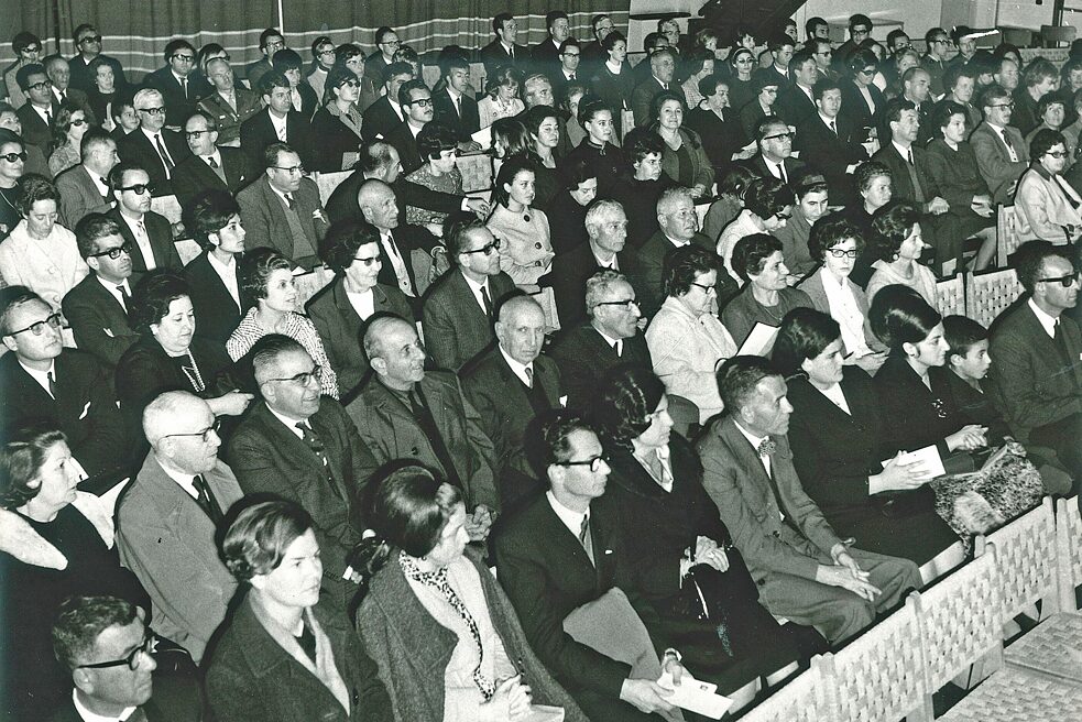 The black and white picture shows the audience at an orchestra concert on 14 February 1969 at the Nicosia School for the Blind. The visitors wear elegant evening attire, women wear dresses and have their hair up. The audience is seated in diagonal rows facing the stage. The first row is empty and shows a white row of chairs.