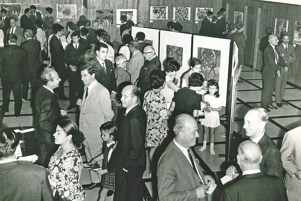 Individual visitors and groups of visitors look at works in the exhibition and talk. The attire is elegant evening dress; there are both children and adults of all ages.