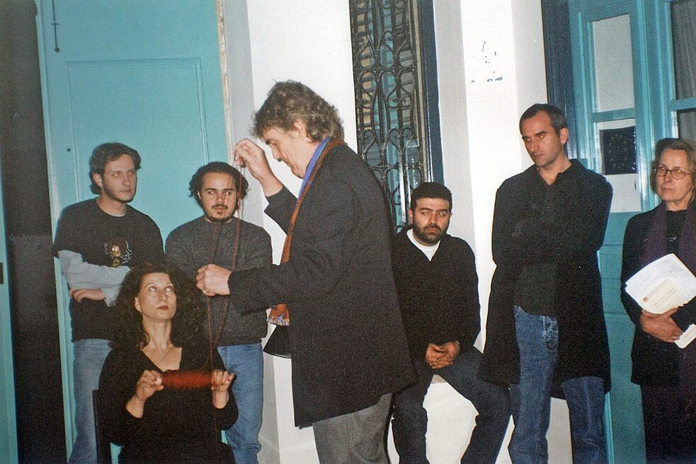 The artist Horst Weierstall presents an abstract installation to an audience. His gaze is fixed on the object in his hands, he is wearing an elegant black suit with brown suit trousers. The six spectators are standing in front of a turquoise door and a window with a turquoise frame; they are looking at the artist intently.