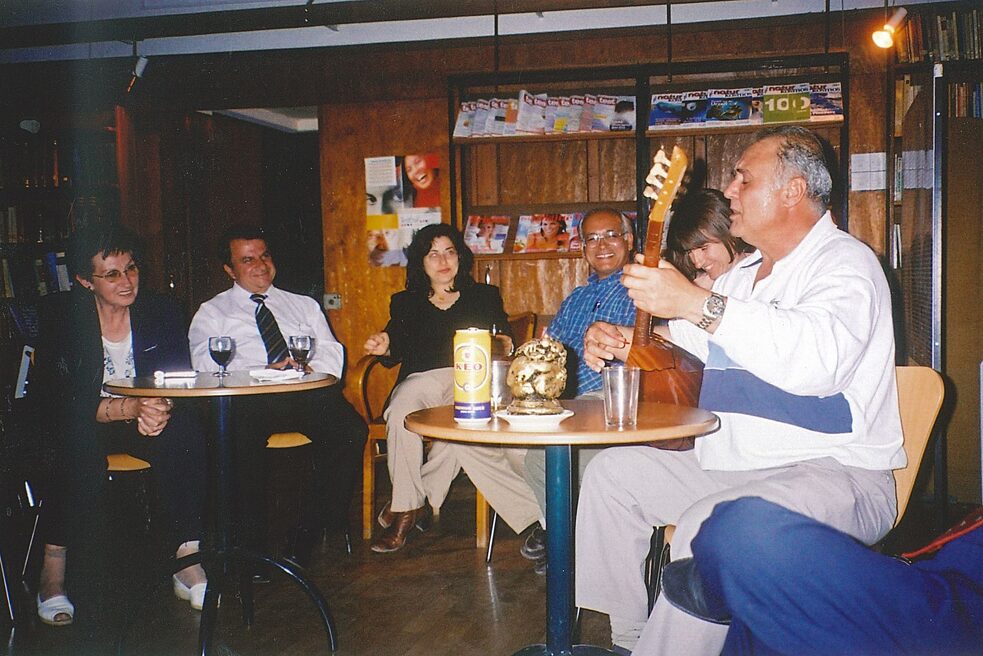 At the event "Meeting Point Green Line" on 4 May shortly after the opening of the checkpoint at Ledra Palace, Emir Gül plays the saz. He wears a white shirt, light grey trousers and sits at a table with a few men and women. On the table are alcoholic drinks such as beer and wine, and in the background on the wall is a shelf with flyers.