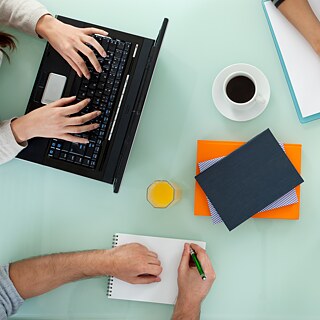Conferentietafel van bovenaf: twee handen aan elk van twee laptops, twee handen met pen over twee notitieblokken, drankjes 