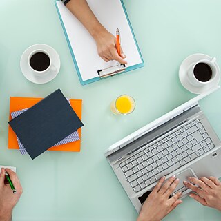 Draufsicht auf einen Tisch mit Laptops, Notizblocks, Schreibgeräten und Kaffeetassen