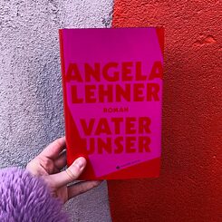 Eine Hand hält Angela Lehners Roman "Vater unser" vor einer grauen Hauswand.