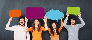 Vier junge Menschen stehen lächelnd vor einem dunklen Hintergrund und halten bunte Sprechblasen über ihren Köpfen. 