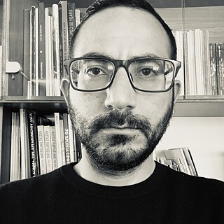 Ein Schwarz-Weiß-Halbporträt eines jungen Mannes mit schwarzen Haaren und Bart, einer quadratischen Brille und einem dunklen T-Shirt. Er starrt mit einem ernsten Blick in die Kamera. Hinter ihm stehen Bücher in einem Regal.