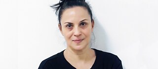 Μισό πορτραίτο μιας γυναίκας με μαύρα μαλλιά πιασμένα σε κότσο και καστανά μάτια. Φορά μια μαύρη μπλούζα με κεντημένα μανίκια, στέκεται μπροστά από έναν λευκό τοίχο και κοιτάζει την κάμερα.