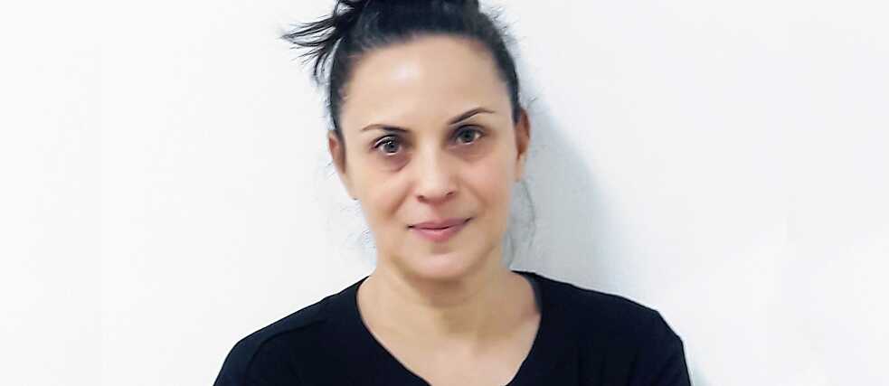 Μισό πορτραίτο μιας γυναίκας με μαύρα μαλλιά πιασμένα σε κότσο και καστανά μάτια. Φορά μια μαύρη μπλούζα με κεντημένα μανίκια, στέκεται μπροστά από έναν λευκό τοίχο και κοιτάζει την κάμερα.