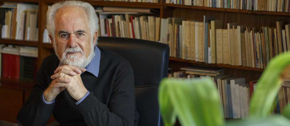 Ένας άνδρας κάθεται σε μια δερμάτινη καρέκλα μπροστά από ένα γραφείο. Έχει άσπρα μαλλιά και άσπρα γένια, φοράει πουλόβερ πάνω από ένα άσπρο και μπλε ριγέ πουκάμισο και έχει μπλεγμένα τα δάκτυλα των χεριών του μπροστά από το πηγούνι του. Στο γραφείο υπάρχει ένα ανοιχτό σημειωματάριο, ένας εκτυπωτής και ένα θολό φυτό, με φόντο ράφια με βιβλία.