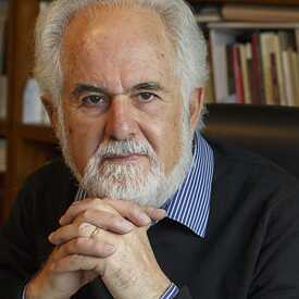 Ένας άνδρας κάθεται σε μια δερμάτινη καρέκλα μπροστά από ένα γραφείο. Έχει άσπρα μαλλιά και άσπρα γένια, φοράει πουλόβερ πάνω από ένα άσπρο και μπλε ριγέ πουκάμισο και έχει μπλεγμένα τα δάκτυλα των χεριών του μπροστά από το πηγούνι του. Στο γραφείο υπάρχει ένα ανοιχτό σημειωματάριο, ένας εκτυπωτής και ένα θολό φυτό, με φόντο ράφια με βιβλία.