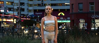 Ein Mädchen im Teenageralter, bekleidet mit silberglänzender Leggings, Bustier und einem Einhorn-Haarreifen, steht bei Dämmerung draußen vor Gebäuden und einer Straße. 