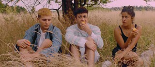 Drei junge Erwachsene sitzen nebeneinander im hohen Gras und essen Früchte. 