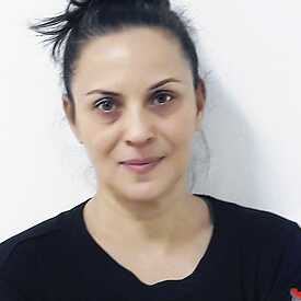 Μισό πορτραίτο μιας γυναίκας με μαύρα μαλλιά πιασμένα σε κότσο και καστανά μάτια. Φορά μια μαύρη μπλούζα με κεντημένα μανίκια, στέκεται μπροστά από έναν λευκό τοίχο και κοιτάζει την κάμερα.
