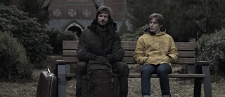 Ein Jugendlicher in einem gelben Regenmantel und ein schwary gekleideter, bärtiger Mann sitzen auf eine Bank