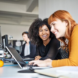 Две насмеани, млади жени седат на биро во канцеларија. Пред нив има повеќе монитори и еден лаптоп.