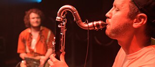 A man in close-up plays the bass clarinet with his eyes closed. In the background, out of focus, a seated man in beige trousers and red shirt holds something in his hands.